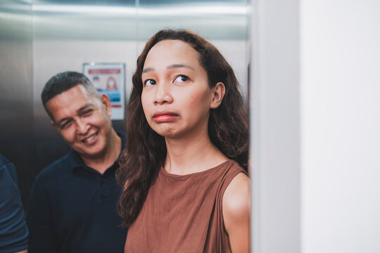 A young asian woman cringes and feels uncomfortable inside an elevator with a creepy man smiling creepily at her. She is wishing for the elevator to open.