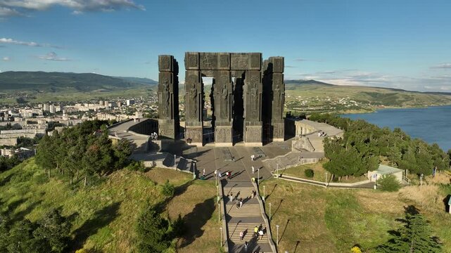 Drone shot capturing the massive bronze and stone pillars of the Chronicle of Georgia monument complex, situated on a green hill overlooking the vast Tbilisi Sea reservoir under a clear blue sky