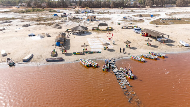 Aerial view of Lake Retba with colorful wooden boats on the pink water, sandy shore with small huts, a heart-shaped sculpture, and people in Rufisque, Dakar Region, Senegal.