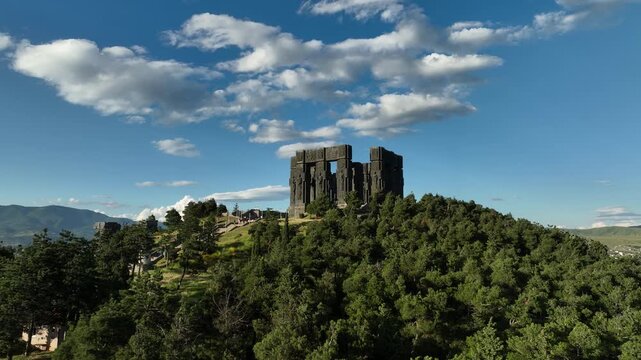Drone shot capturing the massive bronze and stone pillars of the Chronicle of Georgia monument complex, situated on a green hill overlooking the vast Tbilisi Sea reservoir under a clear blue sky
