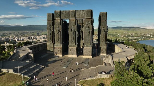 Drone shot capturing the massive bronze and stone pillars of the Chronicle of Georgia monument complex, situated on a green hill overlooking the vast Tbilisi Sea reservoir under a clear blue sky