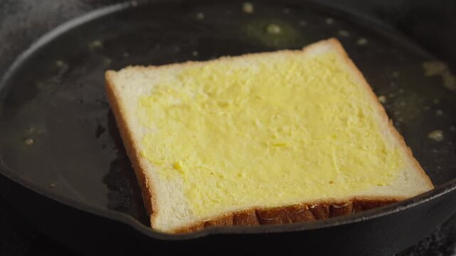 Close up video of a slice of buttered white bread toasting on a hot cast iron skillet