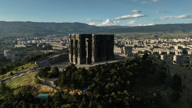 Drone shot capturing the massive bronze and stone pillars of the Chronicle of Georgia monument complex, situated on a green hill overlooking the vast Tbilisi Sea reservoir under a clear blue sky