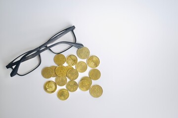 Minimalist flat lay composition showing black spectacles alongside a scattered cluster of golden Malaysian coins on an expansive white background with copy space. © Akram