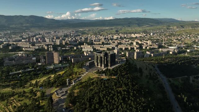 Drone shot capturing the massive bronze and stone pillars of the Chronicle of Georgia monument complex, situated on a green hill overlooking the vast Tbilisi Sea reservoir under a clear blue sky