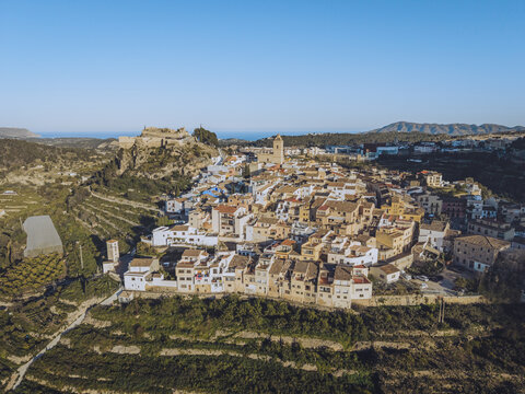 Aerial view of the historic town of Polop with traditional white houses, stone castle ruins, and terraced agricultural fields in Barony of Polop, Valencian Community, Spain.