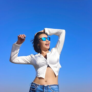 Joyful young Asian woman in a white crop shirt and denim shorts posing with arms raised against a clear blue sky background
