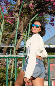Low angle view of a beautiful young Asian woman in denim shorts and a white shirt looking over her shoulder by a fence with pink flower