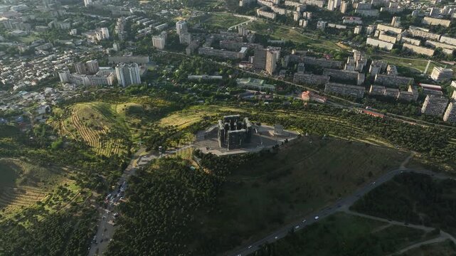 Drone shot capturing the massive bronze and stone pillars of the Chronicle of Georgia monument complex, situated on a green hill overlooking the vast Tbilisi Sea reservoir under a clear blue sky