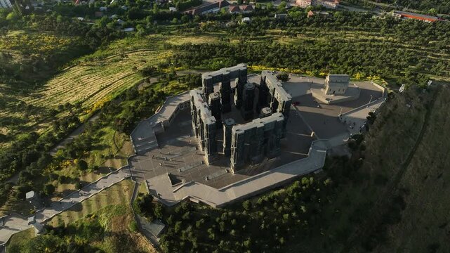 Drone shot capturing the massive bronze and stone pillars of the Chronicle of Georgia monument complex, situated on a green hill overlooking the vast Tbilisi Sea reservoir under a clear blue sky