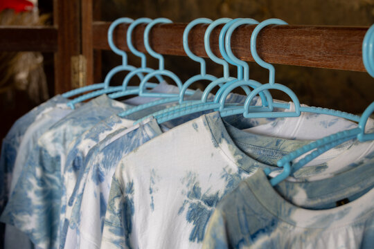 close-up shot of tie-dye t-shirts hanging on light blue plastic hangers. The shirts feature a soft, pastel tie-dye pattern.