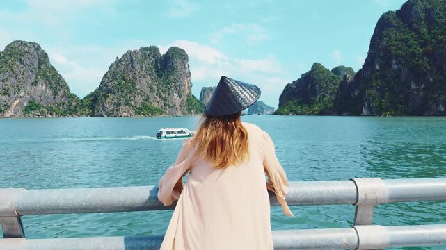 Woman wearing traditional non la hat standing on a viewpoint railing, looking at a tourist boat moving through the calm blue waters of halong bay with lush green limestone karsts under a clear sky