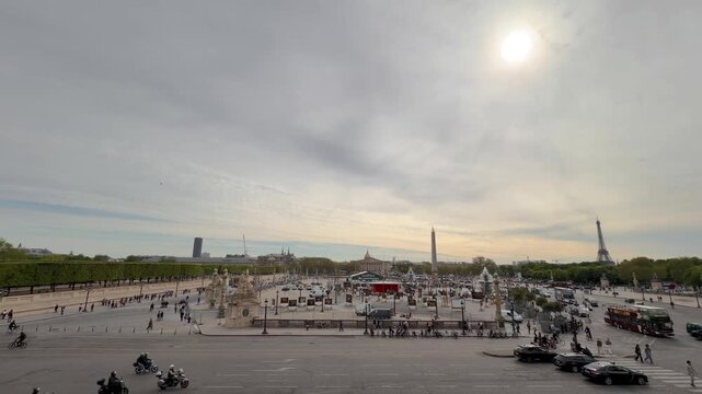 Panoramic view of Place de la Concorde with the Luxor Obelisk, traffic, and tourists, with the Eiffel Tower and Montparnasse Tower visible on the distant skyline against a bright, cloudy sky