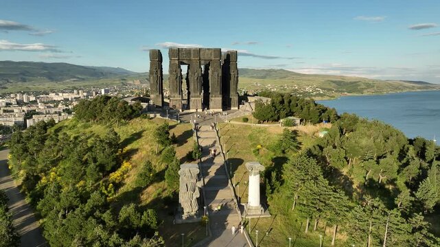 Drone shot capturing the massive bronze and stone pillars of the Chronicle of Georgia monument complex, situated on a green hill overlooking the vast Tbilisi Sea reservoir under a clear blue sky