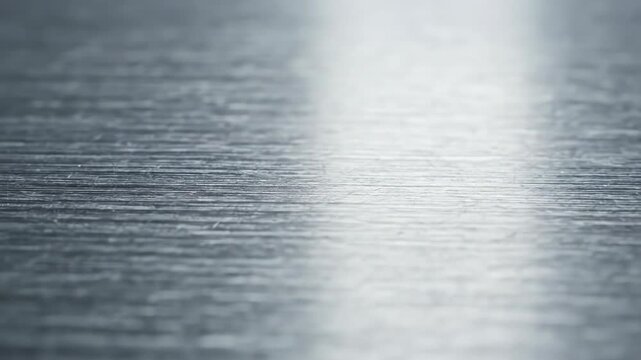 Horizontal brushed silver metal texture macro with reflective light highlights and shallow depth of field focusing on fine metallic grain and linear steel surface pattern.
