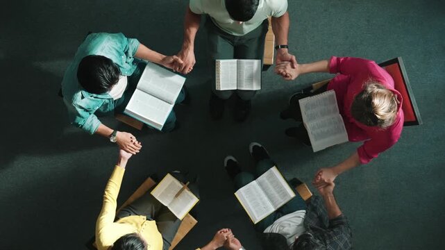 Top down view of diverse people holding hand and sitting in circle with bible book on lap. Aerial view of faithful prayer clasp hand and praying to god with faith, religion conceptional. Symposium.