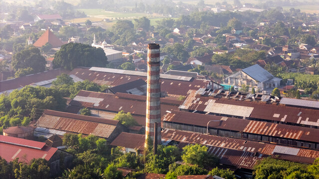 Aerial view of an old sugar factory with a tall striped chimney and rusted metal rooftops, surrounded by a dense residential town and lush green trees in Indonesia.