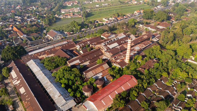 Aerial view of an old industrial factory complex with a tall brick chimney and weathered metal roofs surrounded by lush green trees and rural landscape in Indonesia.