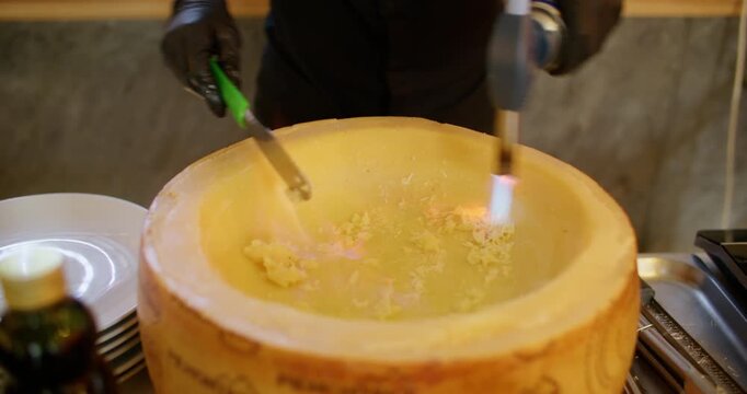 Gourmet cooking. Melting cheese. Chef hand heating grated parmesan wheel piece with kitchen torch flame stirring ingredient with scraper.