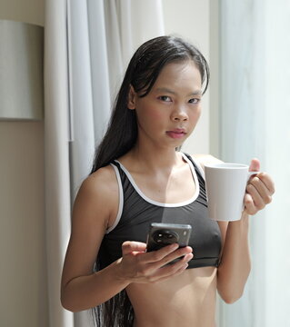 Young Asian woman wearing a black sports bra and shorts stands near a window while holding a white coffee mug and a smartphone