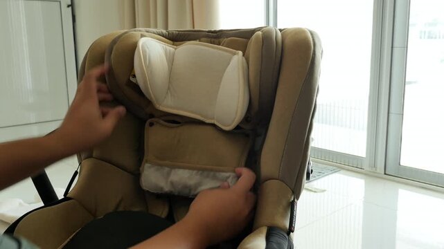 A man adjusts the headrest and secures the straps on a child car seat near a large window. Suitable for safety tutorials or parenting guides.