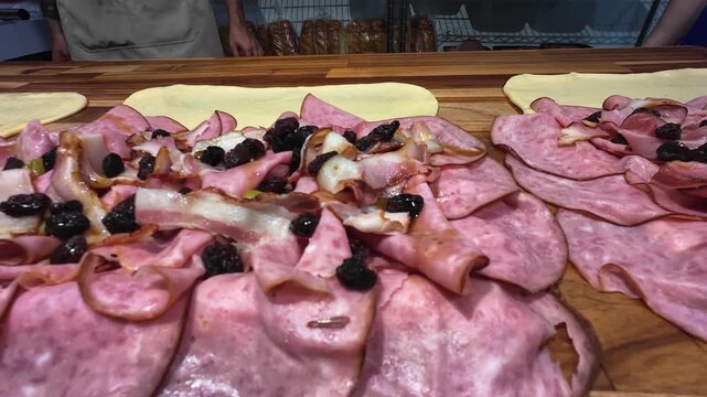 Close-up sliced Italian deli meats arranged on a wooden preparation table with olives and fresh ingredients. Artisanal food preparation, charcuterie with detailed textures and authentic gastronomy