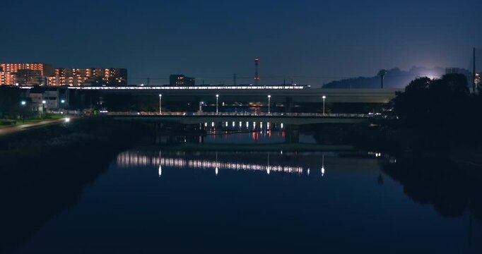 High speed passenger train traveling at night on elevated railway over river. Urban lights reflecting on water. Modern city suburb. Wide static shot, public transportation system, urban Japan mobility