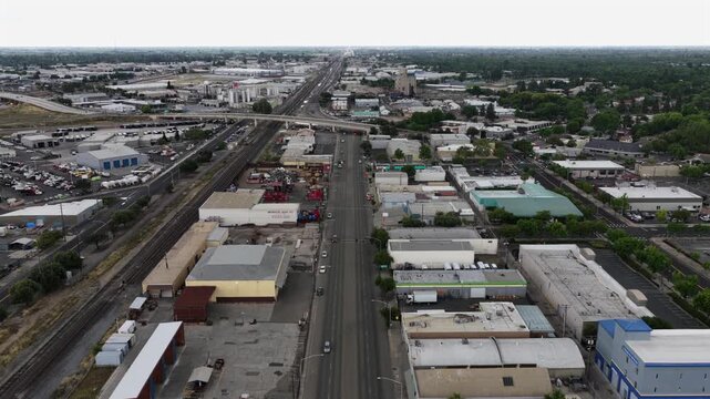 Industrial corridor with warehouses, rail yard, flat commercial rooftops and wide straight road stretching toward downtown and tree-lined neighborhoods in Modesto, California, USA.Aerial drone flyover