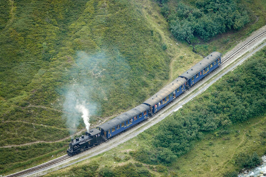 Aerial view of the historic Dampfbahn Furka Bergstrecke steam train traveling through the lush green mountainside in Aarau, Aargau, Switzerland.