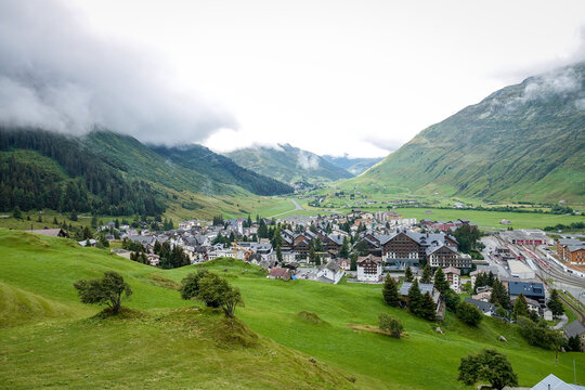 Aerial view of Andermatt village nestled in a lush green valley surrounded by mountains under a cloudy sky in Andermatt, Uri, Switzerland.