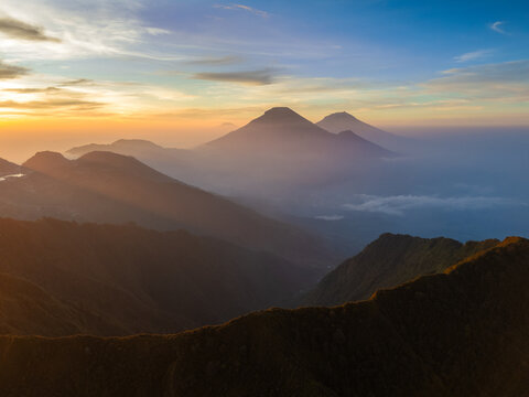 Aerial view of the mountainous landscape near Mount Bismo at sunrise, with golden light illuminating the misty peaks and deep valleys in Kecamatan Kejajar, Jawa Tengah, Indonesia.