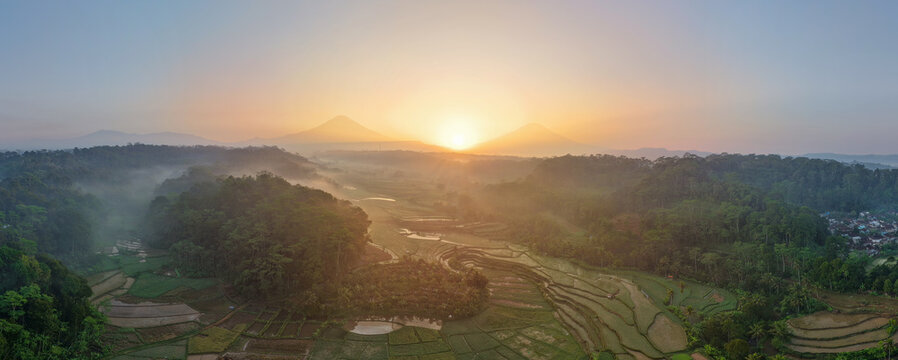 Aerial view of rice terraces nestled among lush hills during a golden sunrise with distant mountains in Selomerto, Central Java, Indonesia.