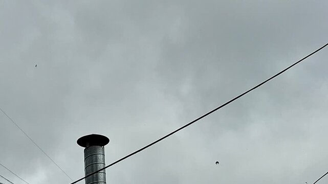 Storm clouds gather above an urban rooftop chimney and power lines, creating a dramatic city skyline scene. Moody weather, shifting light, and atmospheric skies capture the approach of a storm