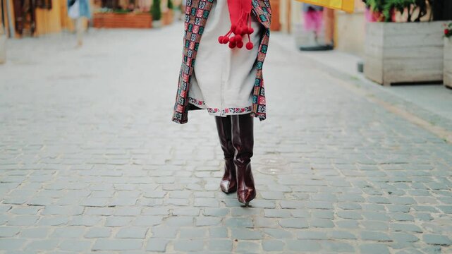 Low-angle close-up of a woman's legs walking on a historic cobblestone street. She is wearing traditional Ukrainian folk clothing, including patterned skirt, white trousers, and burgundy leather boots