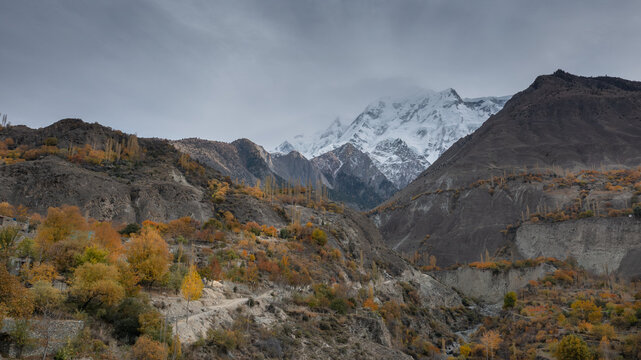 Aerial view of Hunza Valley featuring snow-capped mountain peaks and autumn foliage on rocky slopes under a cloudy sky in Garelt, Gilgit, Pakistan.