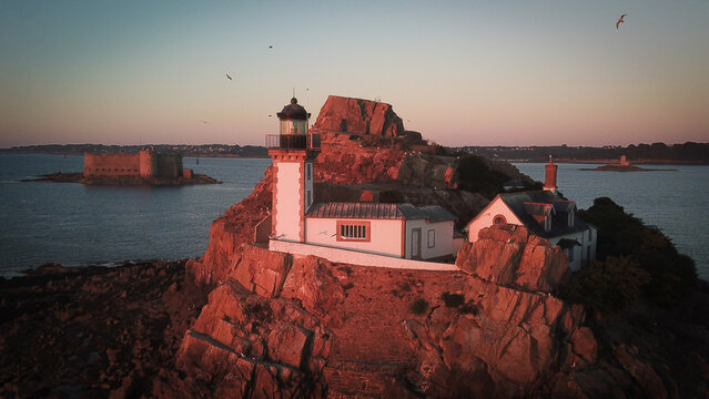 Aerial view of the lighthouse on Louet island at sunset, with rocky terrain, surrounding sea, and the historic Chateau du Taureau in the distance in Carantec, Brittany, France.