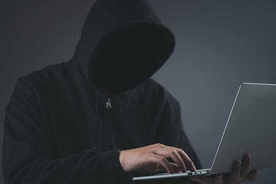 Anonymous hooded hacker using laptop in dark environment, representing cybercrime, data breach, hacking, cybersecurity threat, identity theft, malware attack, digital privacy risk in modern technology