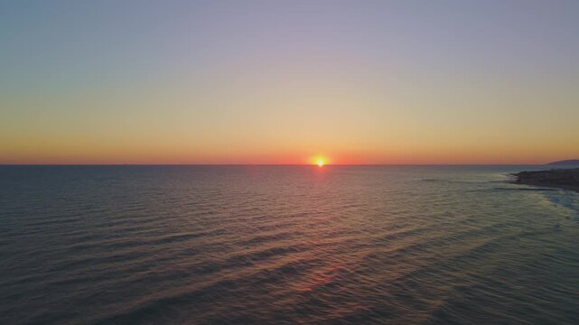 Cinematic drone footage capturing a bright sun rising directly over the clear horizon line of the Mediterranean sea, with soft water ripples and a warm gradient morning sky in Crete, Greece.