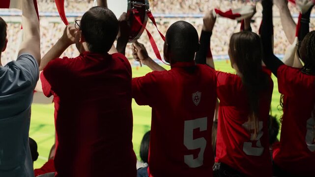 Back view of loud supporter group celebrating dramatic goal during live soccer match in stadium terrace stands, hardcore fans in the supporter section waving scarves and accessories, passion.