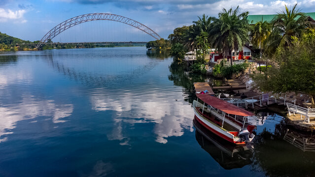 Aerial view of the Adomi Bridge spanning the Volta River with a boat docked at the riverbank, lush palm trees, and reflections of clouds on the water in Atimpoku, Volta Region, Ghana.