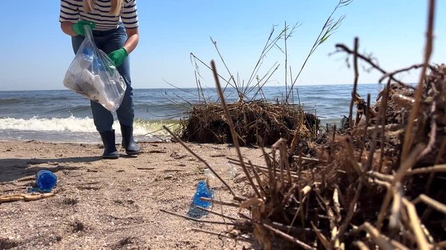 Volunteer bends down to collect plastic bottles and debris from sandy beach. Environmental conservation concept emphasizes the importance of reducing marine pollution and protecting coastlines