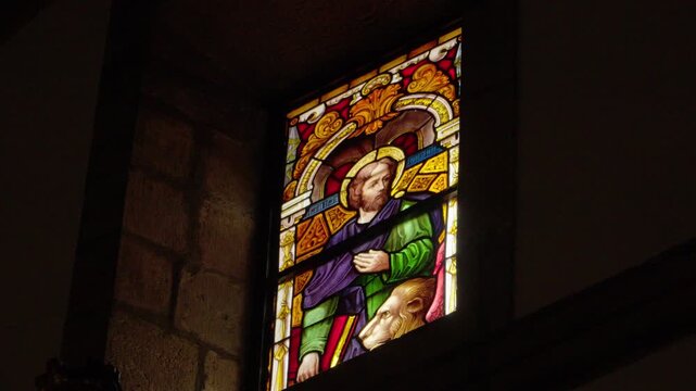 Close-up of illuminated stained glass depicting a saint with halo in a dark stone church interior.