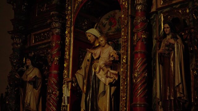 Dimly lit interior shot of ornate gilded altarpiece with Virgin Mary holding infant Jesus, flanked by saints. Ideal for religion, faith, Catholic, and Spanish heritage content.