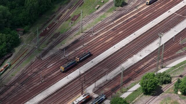 An aerial view captures electric locomotives navigating a complex network of tracks at a large railway station in Hungary.