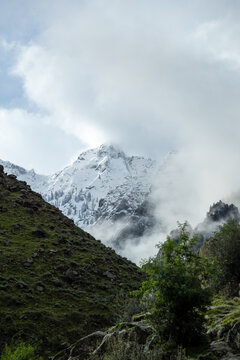 View of the snow-capped Nanga Parbat mountain partially obscured by clouds, rising above a rocky, green-covered slope in Jaglot, Gilgit, Pakistan.