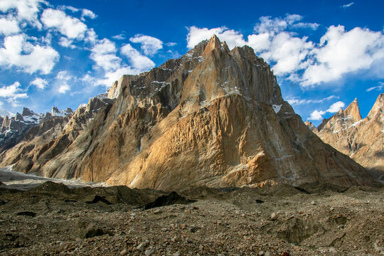 View of the granite spires of the Trango Towers rising above a rocky glacial moraine under a bright blue sky with scattered clouds in the Karakoram range, Pakistan.