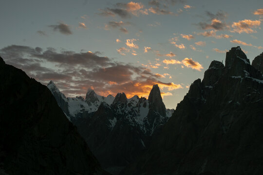View of the jagged granite spires of the Trango Towers in the Karakoram mountain range at sunset, with snow-capped peaks and dramatic clouds illuminated by golden light, Pakistan.