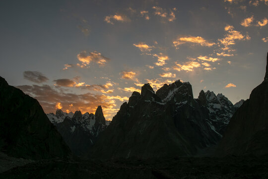 View of the jagged granite spires of the Trango Towers at sunset with golden light illuminating the clouds above the Karakoram mountain range in Pakistan.