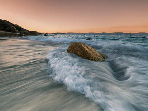 View of the surf washing over a large rock on the sandy beach at sunset in Frenchman Bay, Western Australia, Australia.