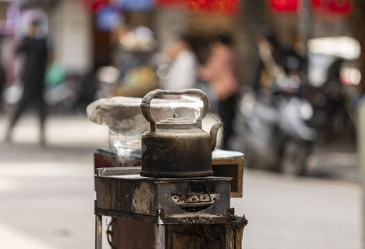 View of a steaming metal kettle resting on a small charcoal stove on a busy street in Hanoi, Ha Noi, Vietnam.